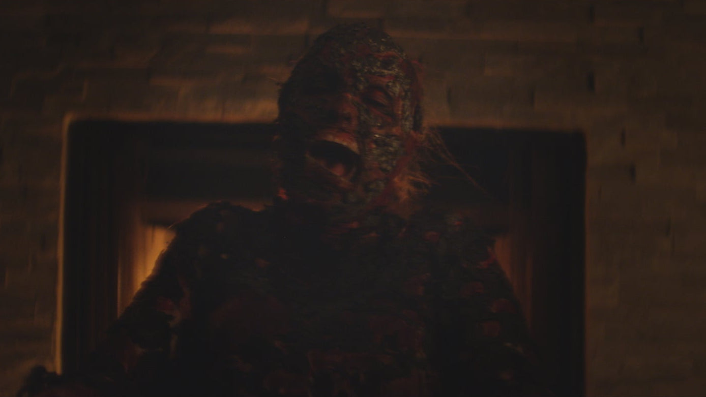 Person wearing a camouflage mask sitting in a dark room with a brick wall.