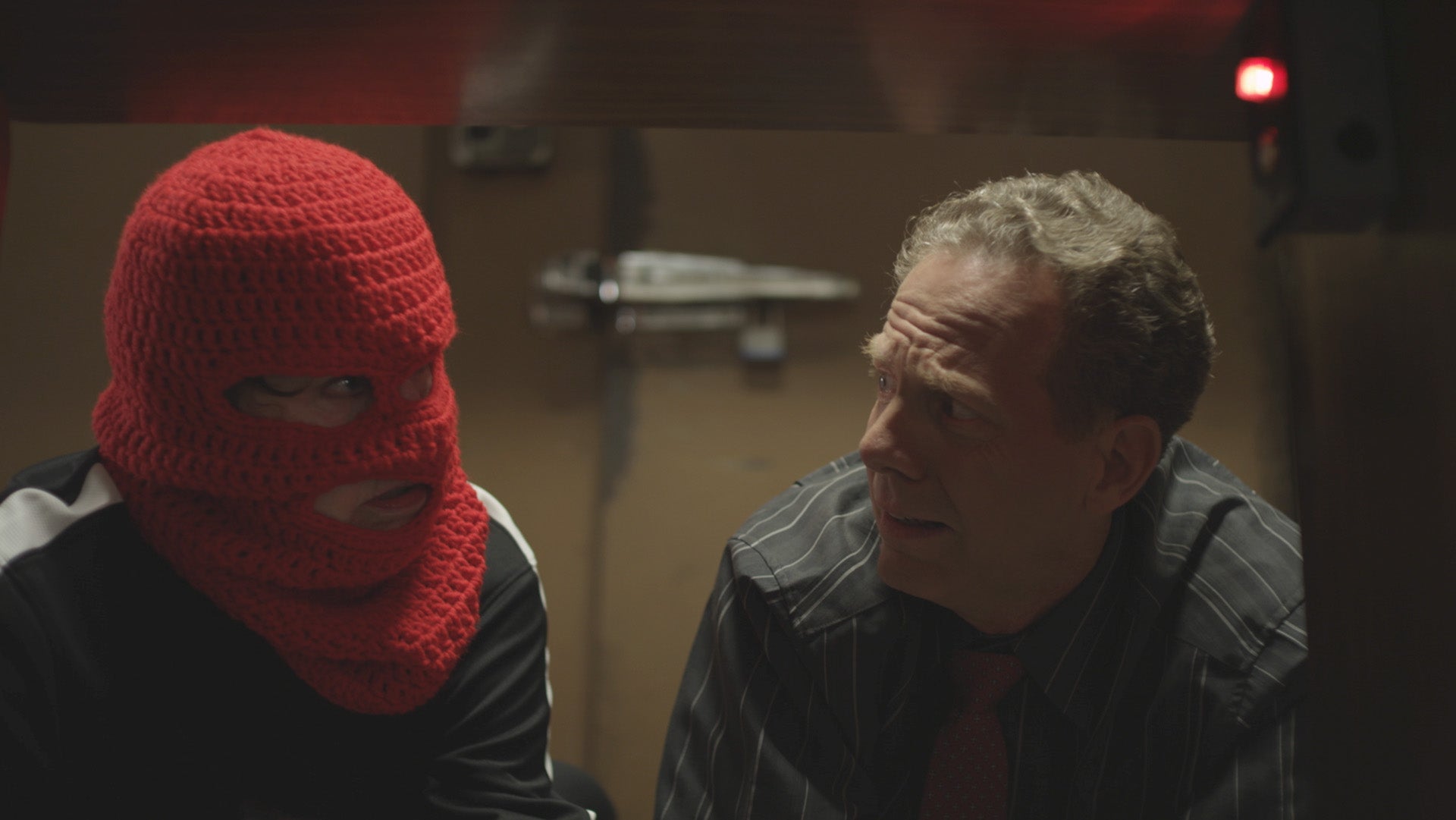Two people in a dimly lit room, one wearing a red knitted balaclava.