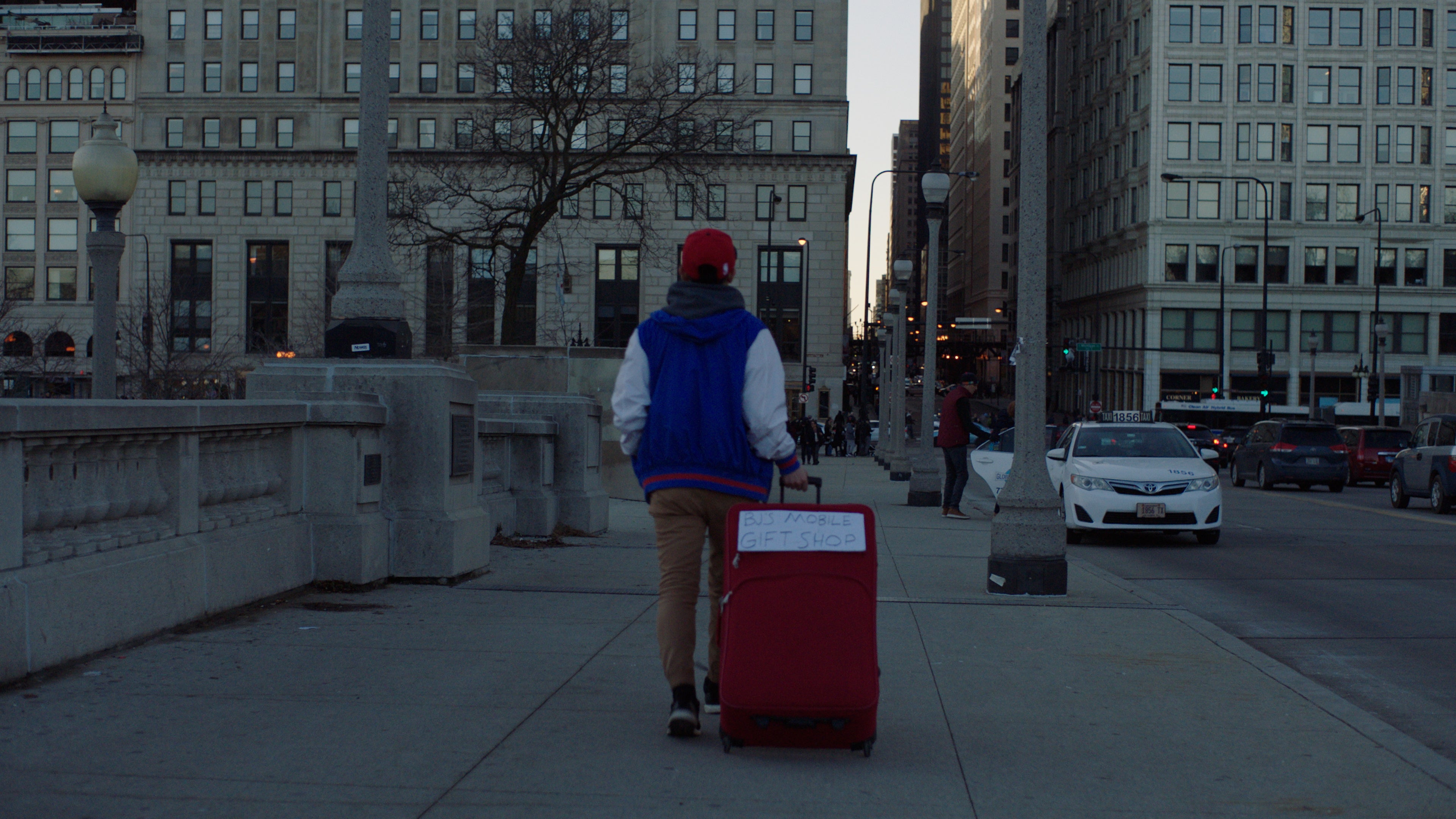 Person with a red suitcase walking on a city street with buildings and cars in the background