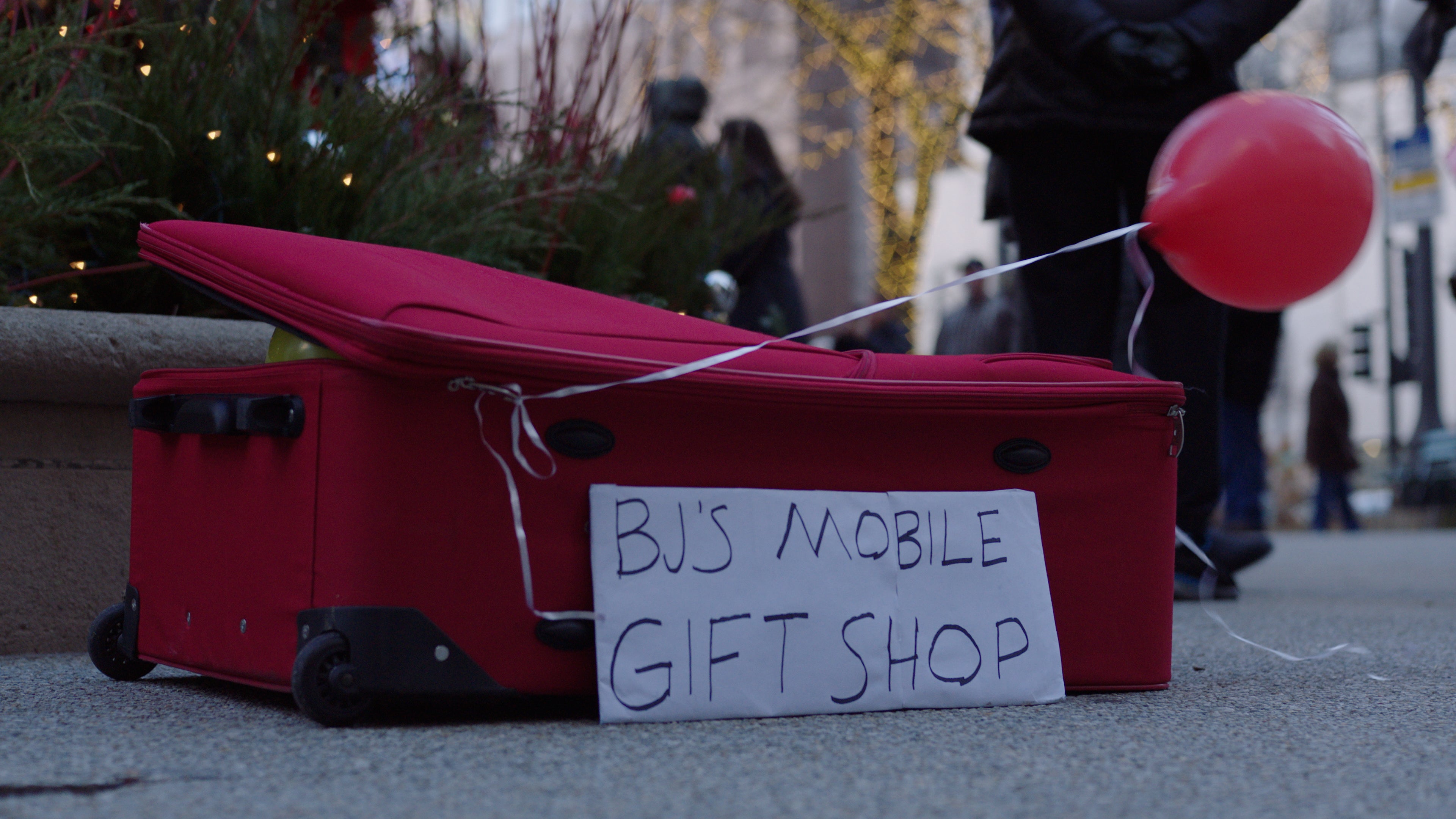 Red suitcase labeled 'BJ's Mobile Gift Shop' on a street with people and festive decorations in the background.