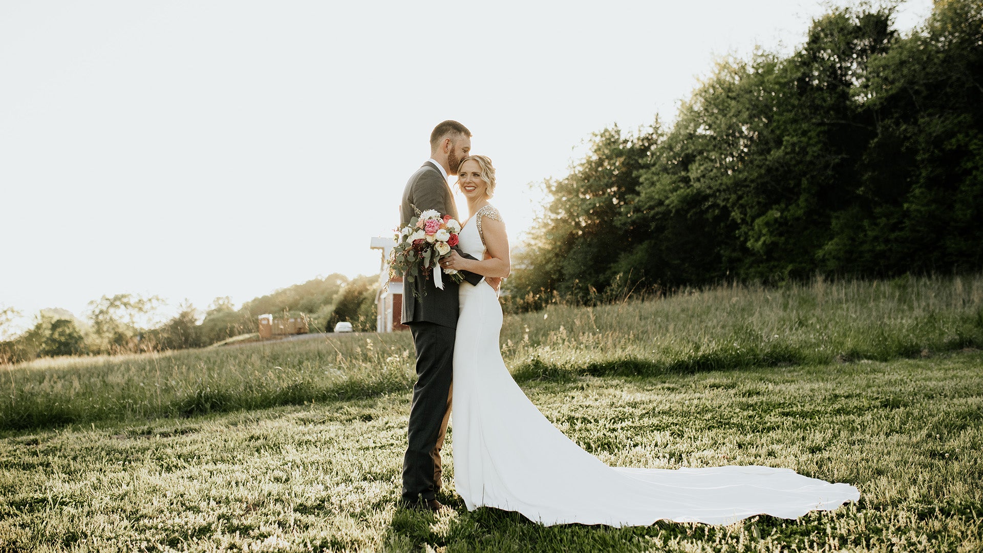 Couple in wedding attire standing in a grassy field with trees in the background