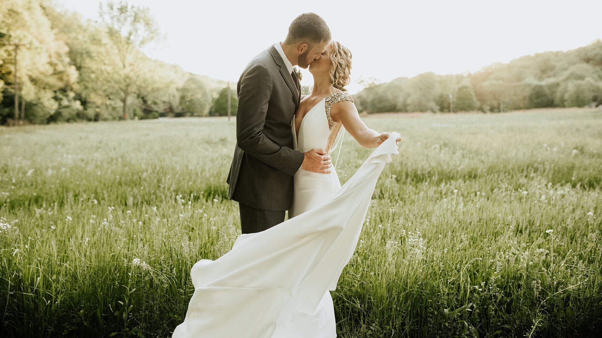 Couple in wedding attire embracing in a field with trees in the background