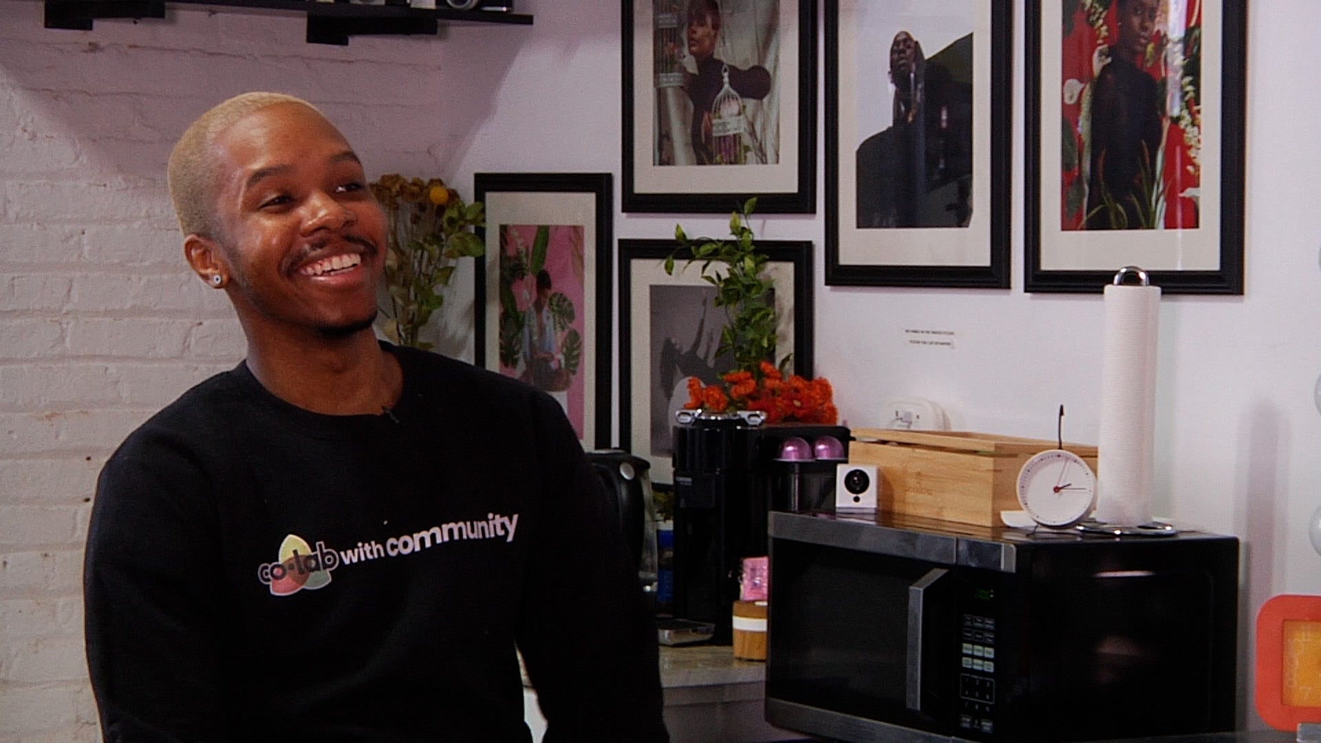 Person wearing a black sweatshirt with text, standing in a room with framed pictures on the wall.