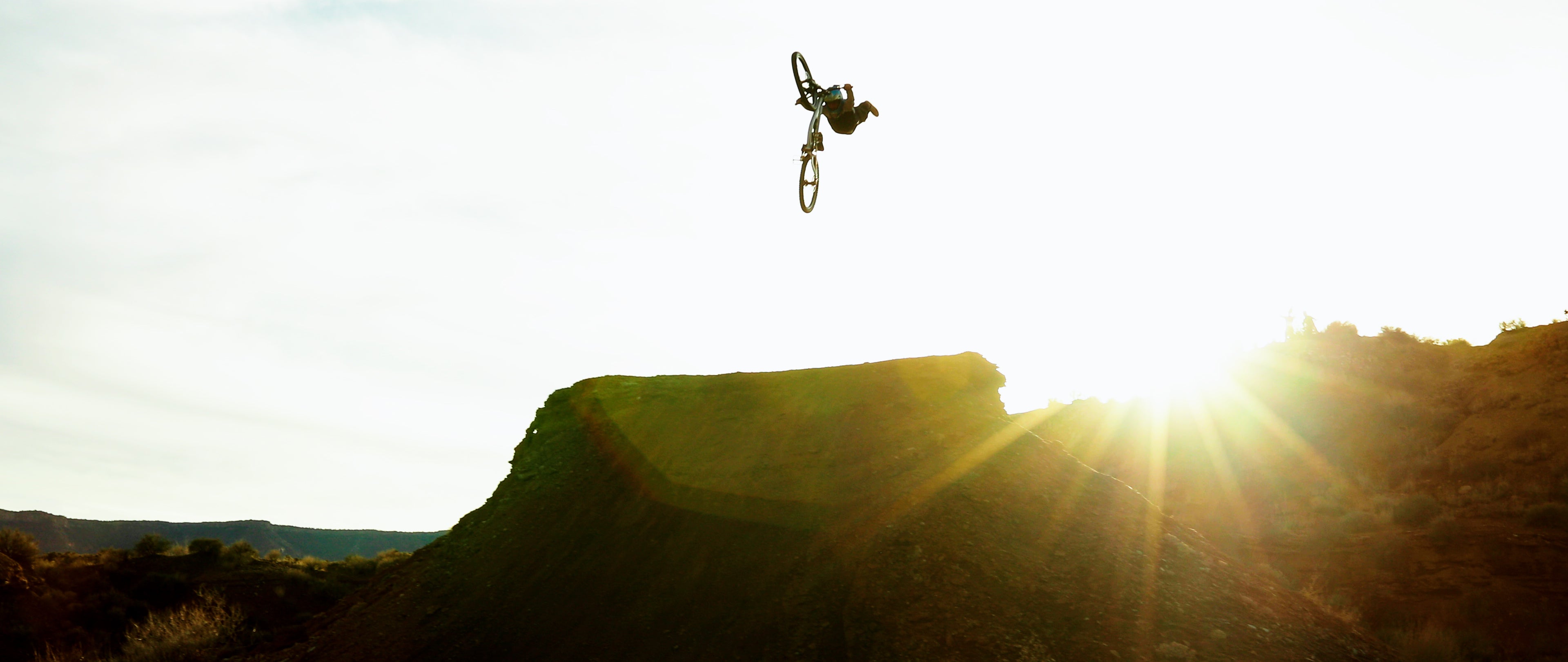 Person on a bicycle performing a jump over a rocky landscape with the sun setting in the background.
