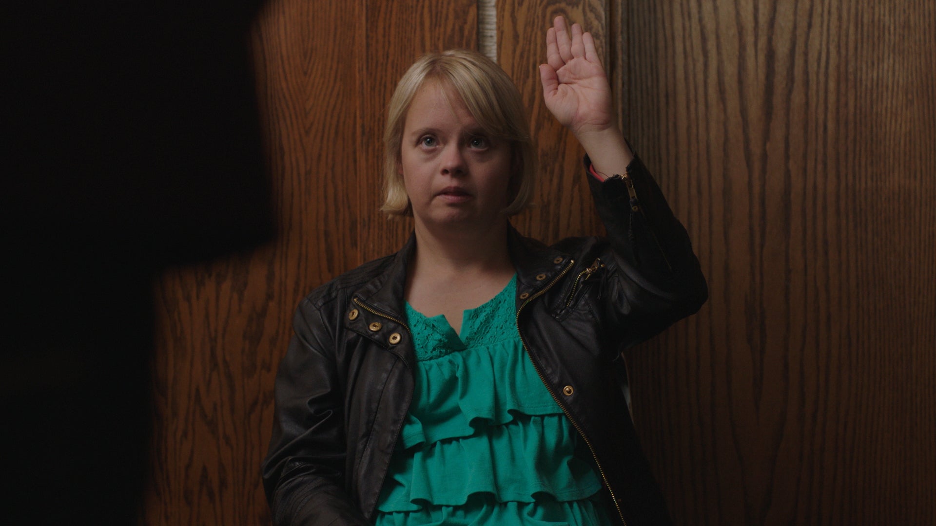Woman in teal blouse and black jacket standing against a wooden wall with one hand raised.