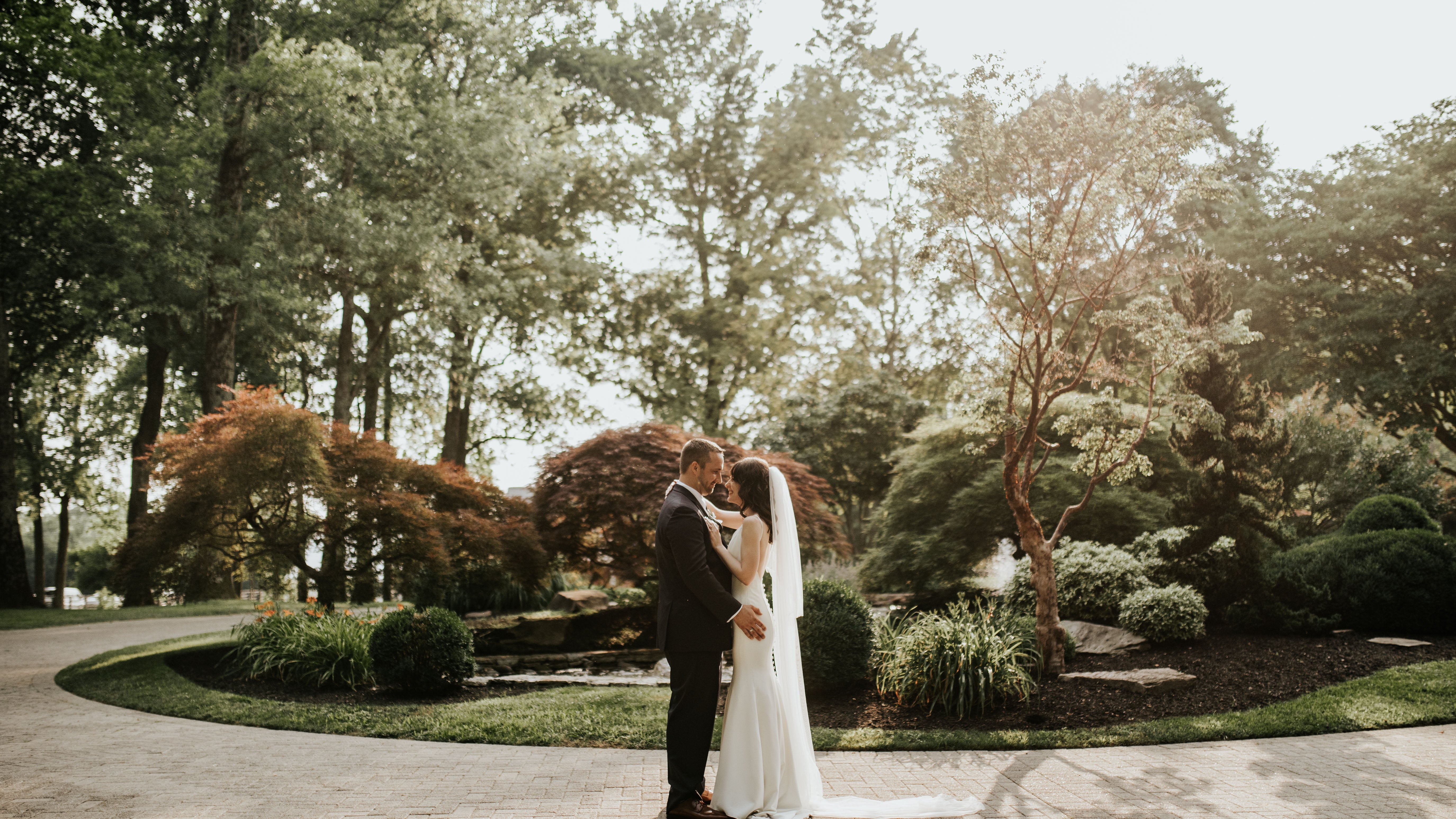 Couple in wedding attire standing in a park with trees and greenery.