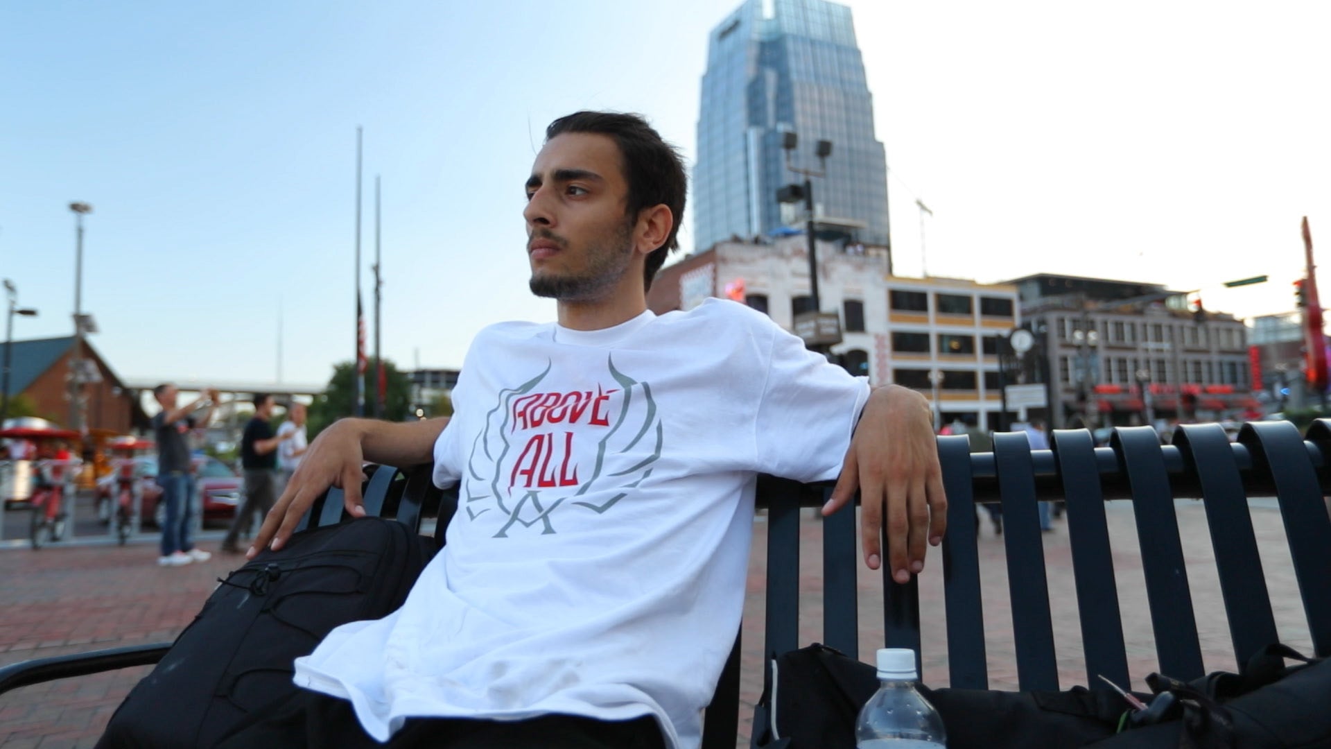 Person wearing a white t-shirt with red text sitting on a bench in an urban setting.
