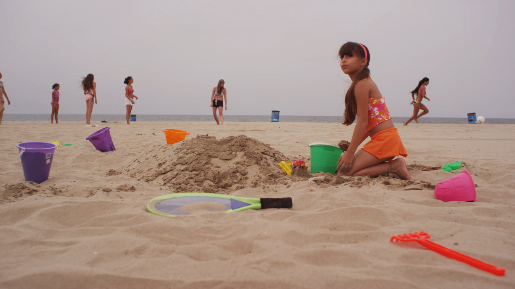 Girl playing in the sand with toys on a beach, other people in the background.