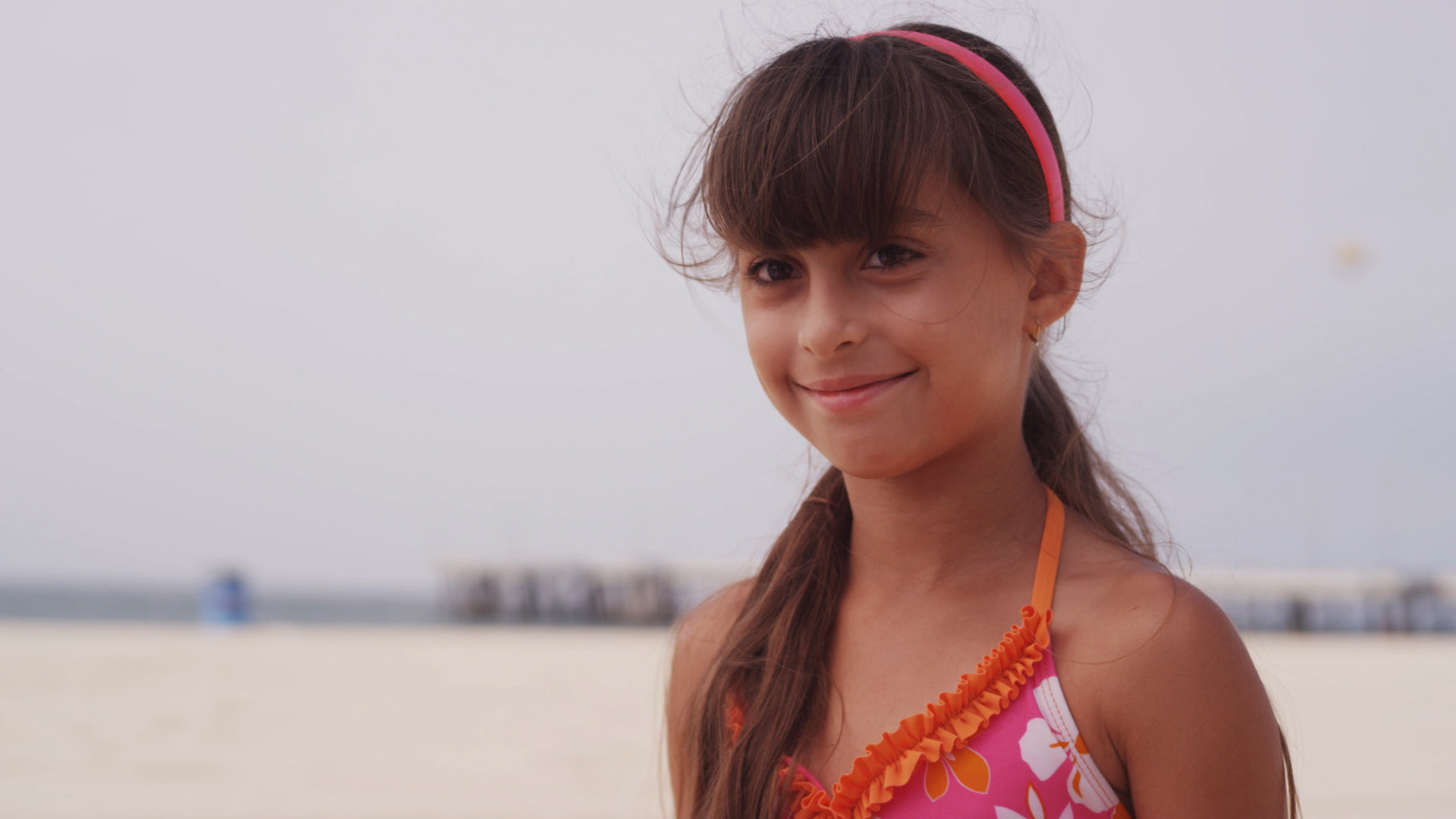 Young girl with a pink headband and orange and pink swimsuit on a beach.
