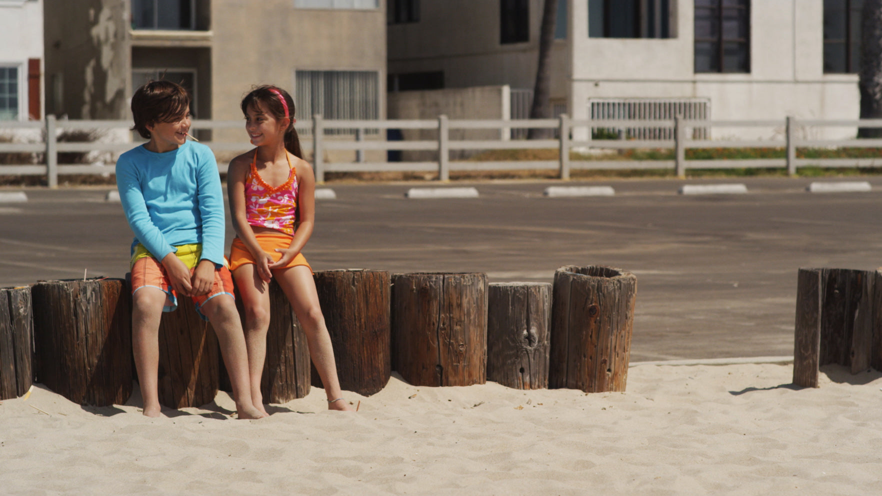 Two children sitting on wooden posts at a beach with a building in the background.