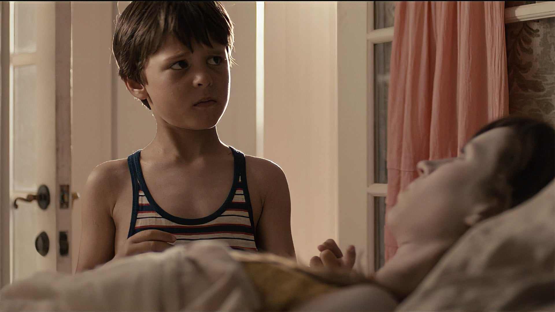 Young boy in a striped tank top sitting on a bed next to a person lying down.