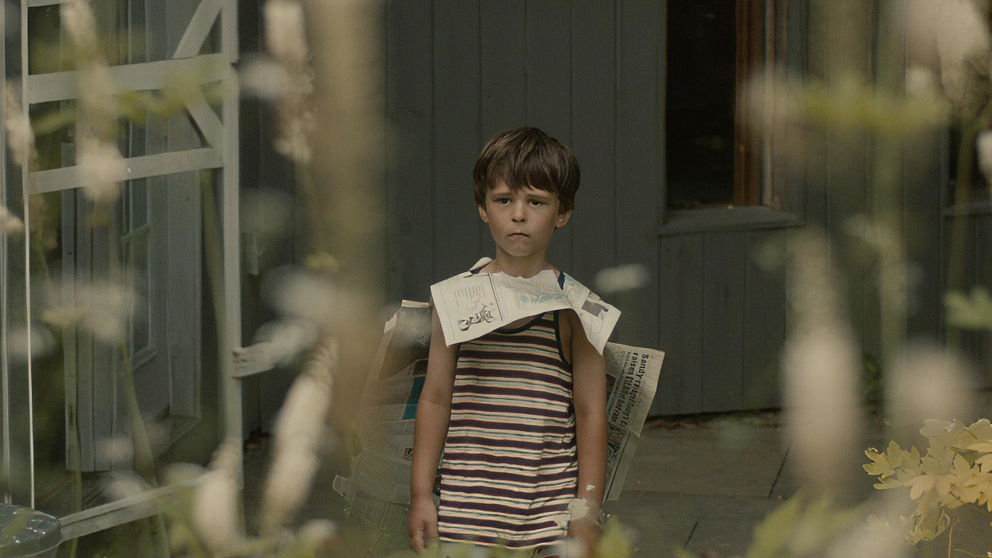 Child wearing a striped dress with newspaper pieces on shoulders in front of a house.