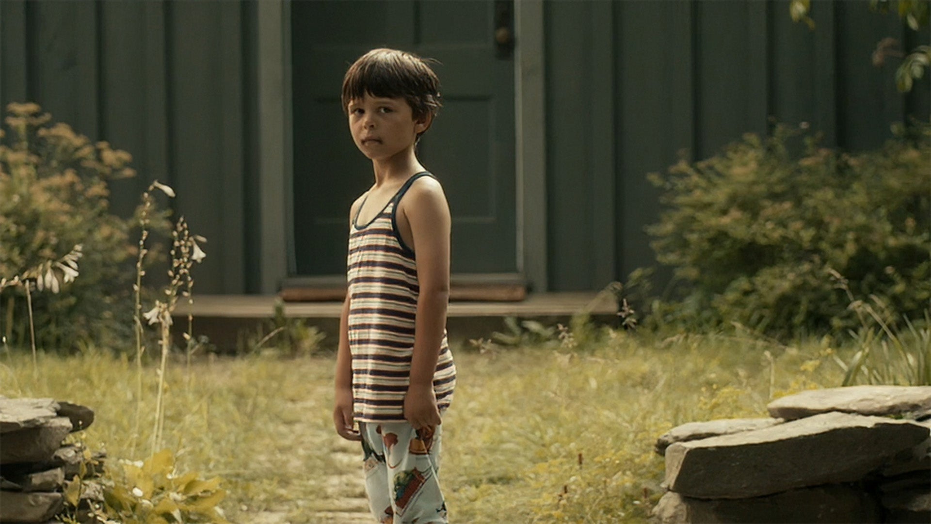 Child in a striped tank top and shorts standing in a grassy area with a building in the background