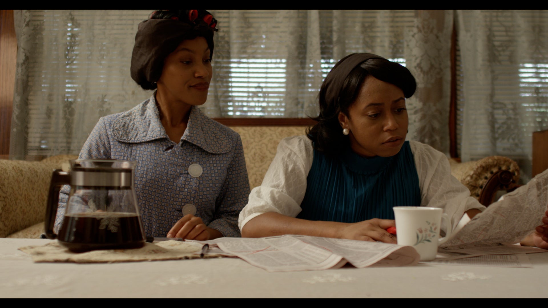 Two women sitting at a table with a coffee pot and newspaper, in a room with curtains.
