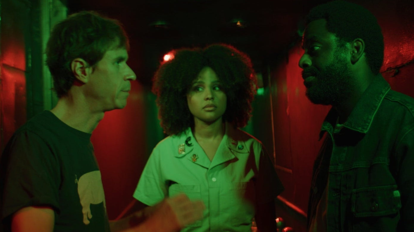 Three people are engaged in a conversation in a dimly lit room with red lighting.