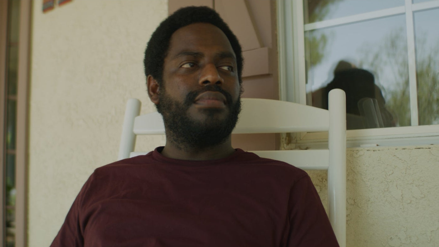 Man sitting on a porch with a guitar, looking off to the side.