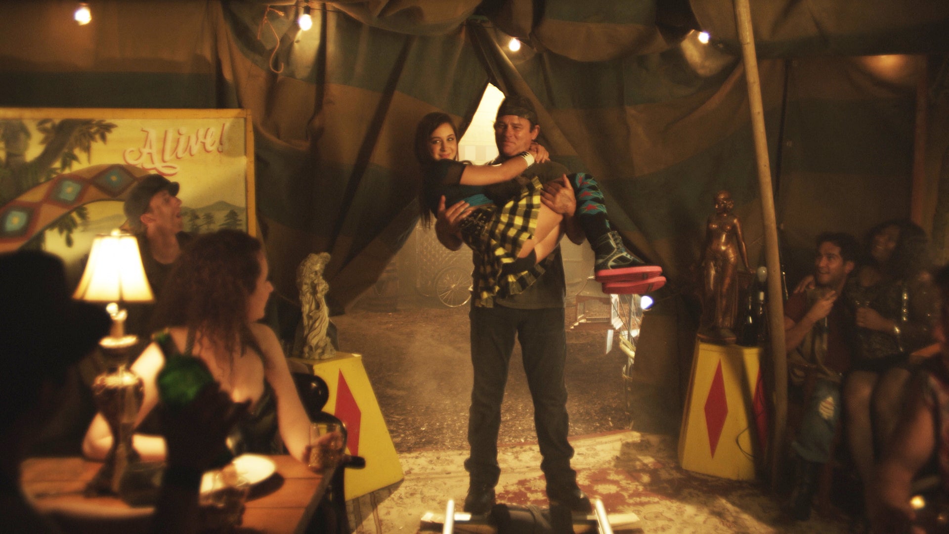 Two people embracing in a dimly lit tent with a vintage circus poster in the background.