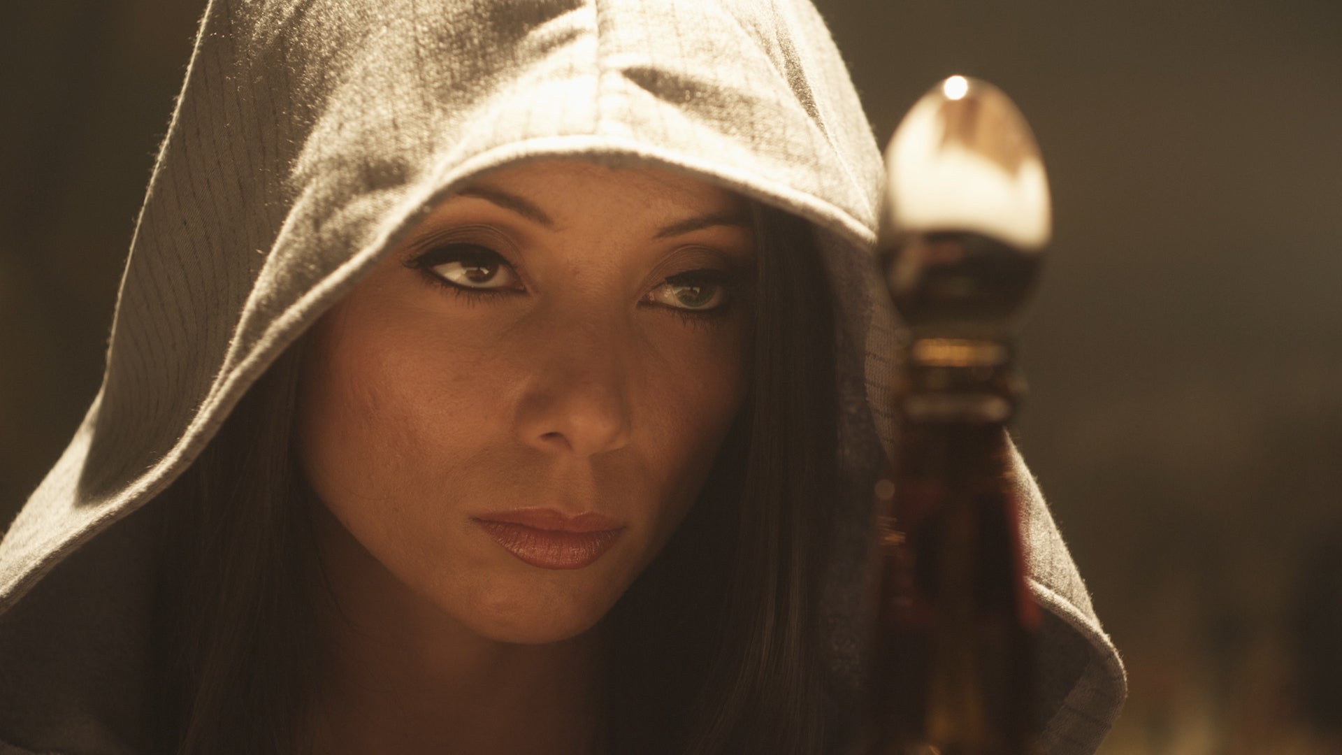 Person in a hooded cloak holding a staff with a glowing top against a dark background