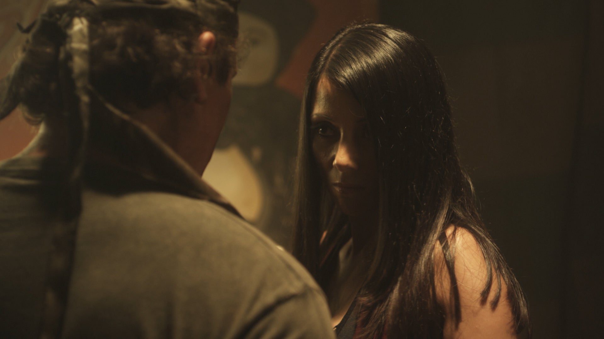 Two people in a dimly lit setting, one facing away from the camera.