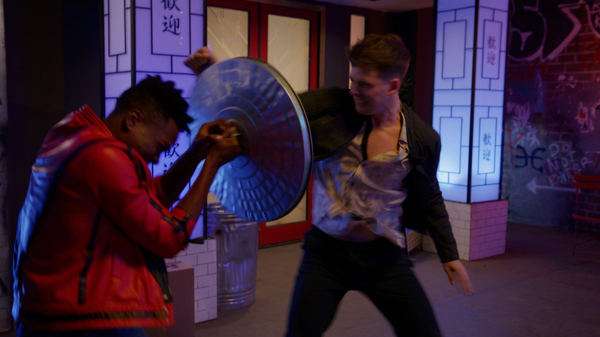 Two people in a room with graffiti and a large blue shield.