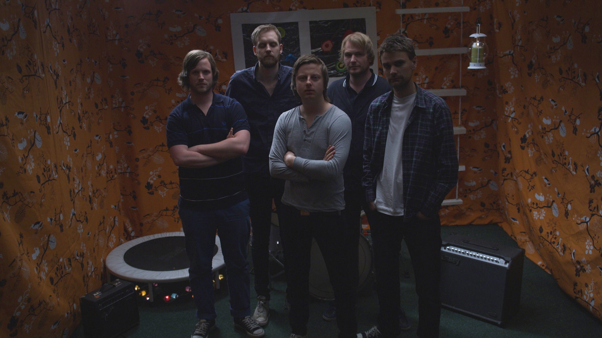 Five men standing in a room with orange walls and musical equipment.