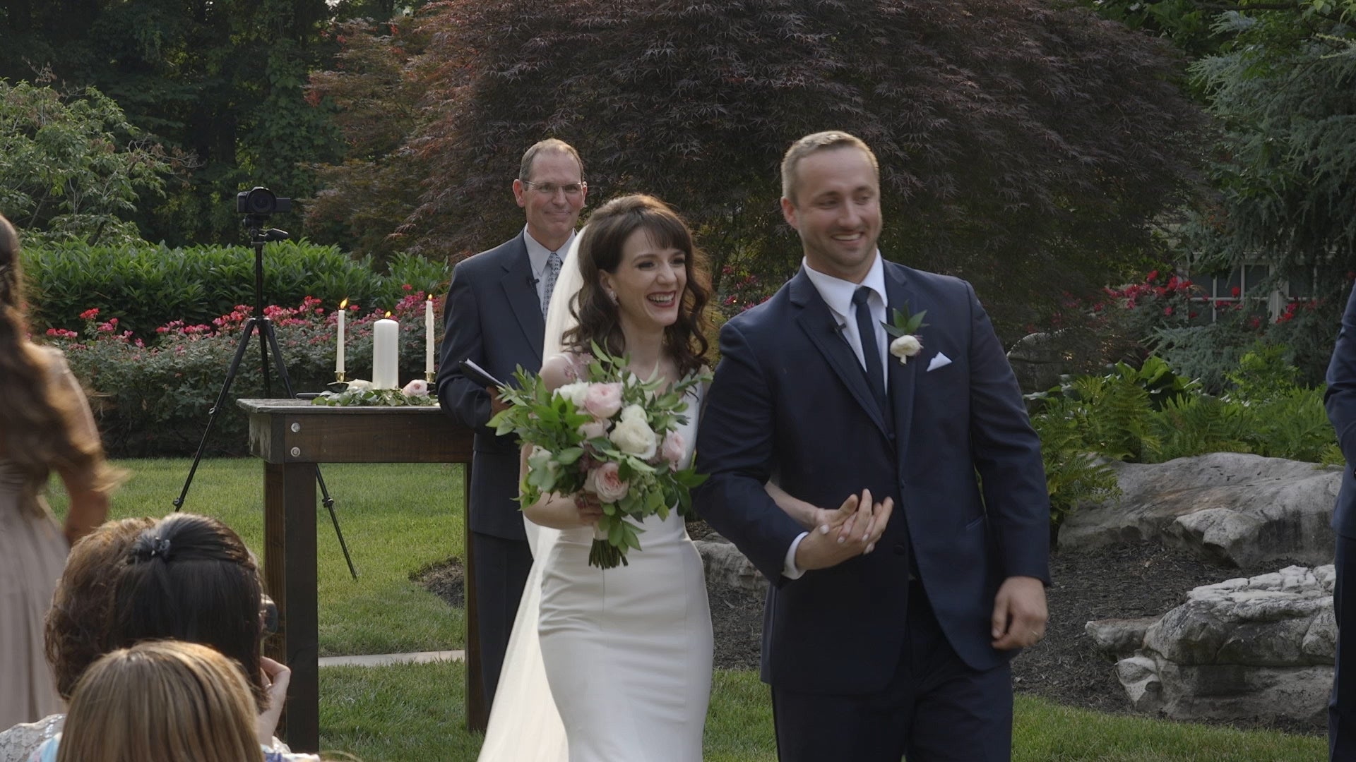 Wedding couple walking outdoors with guests and a floral arrangement on a table.