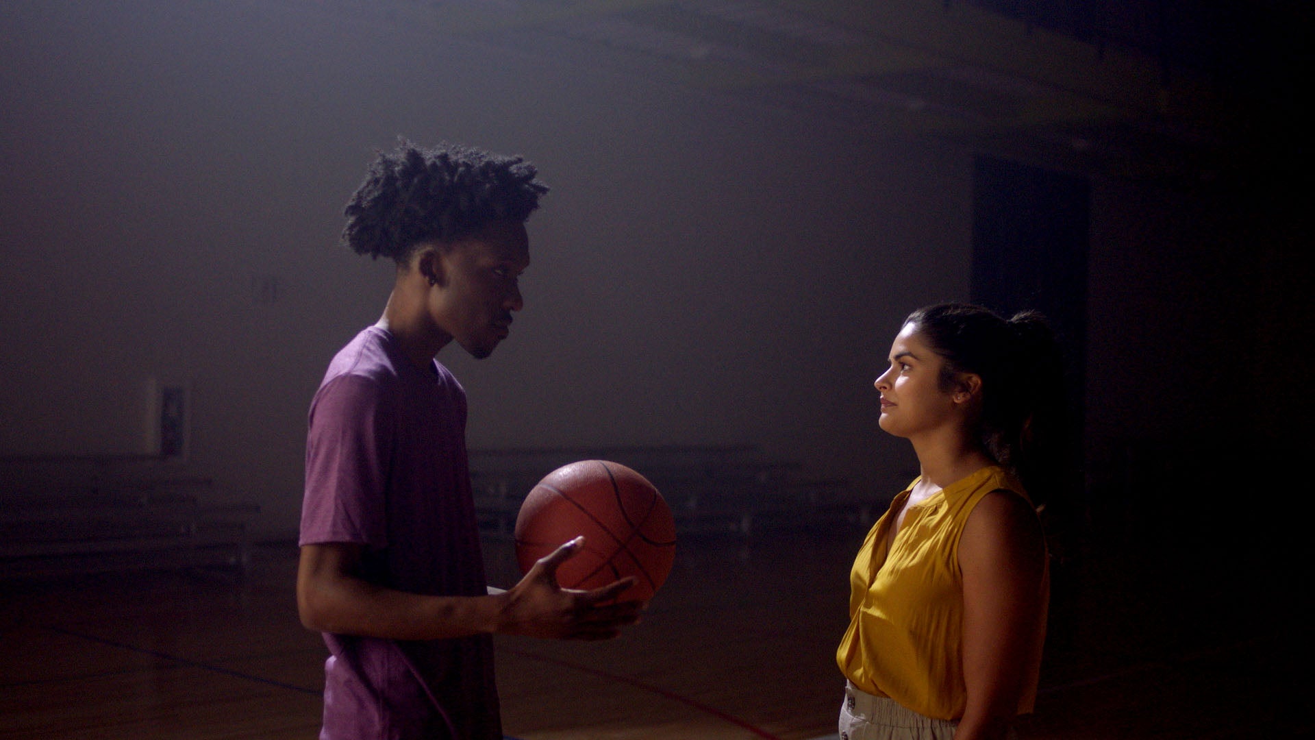 Two people in a gymnasium, one holding a basketball, the other leaning against the wall.