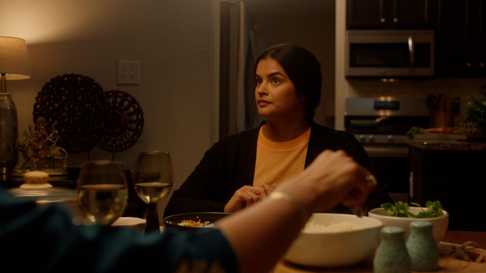 Woman sitting at a dinner table in a home setting with a person partially visible in the foreground.